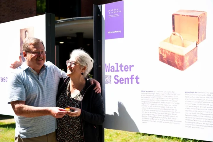 Patrick May and Catherine Guebey-Robert embrace beside an exhibition panel about Walter Senft, holding his wedding ring